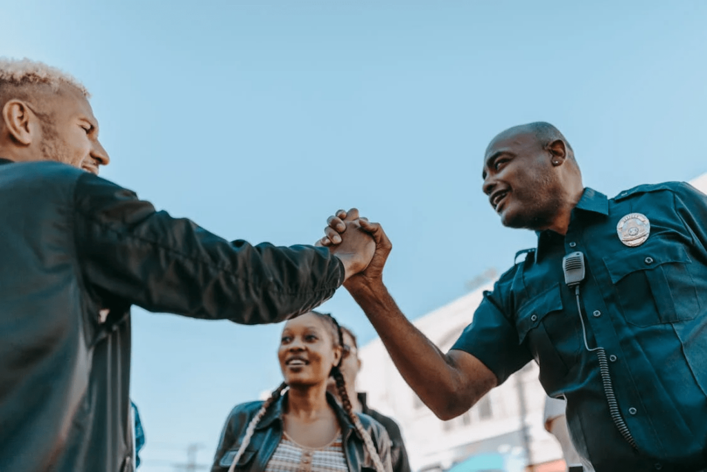 A police officer shakes the hand of a smiling man with several people standing in the background.